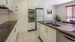 A compact kitchen with white cabinets, a microwave, oven, and refrigerator. There is a sink with a modern faucet, a dining area with a table and chairs, and a window with blinds. - Annand Mews Serviced Apartments (Harristown)