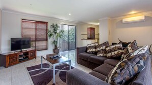 A modern living room with a flat-screen TV, a coffee table, a sofa with patterned cushions, a potted plant, and a sliding glass door leading to a balcony. - Annand Mews Serviced Apartments (Harristown)