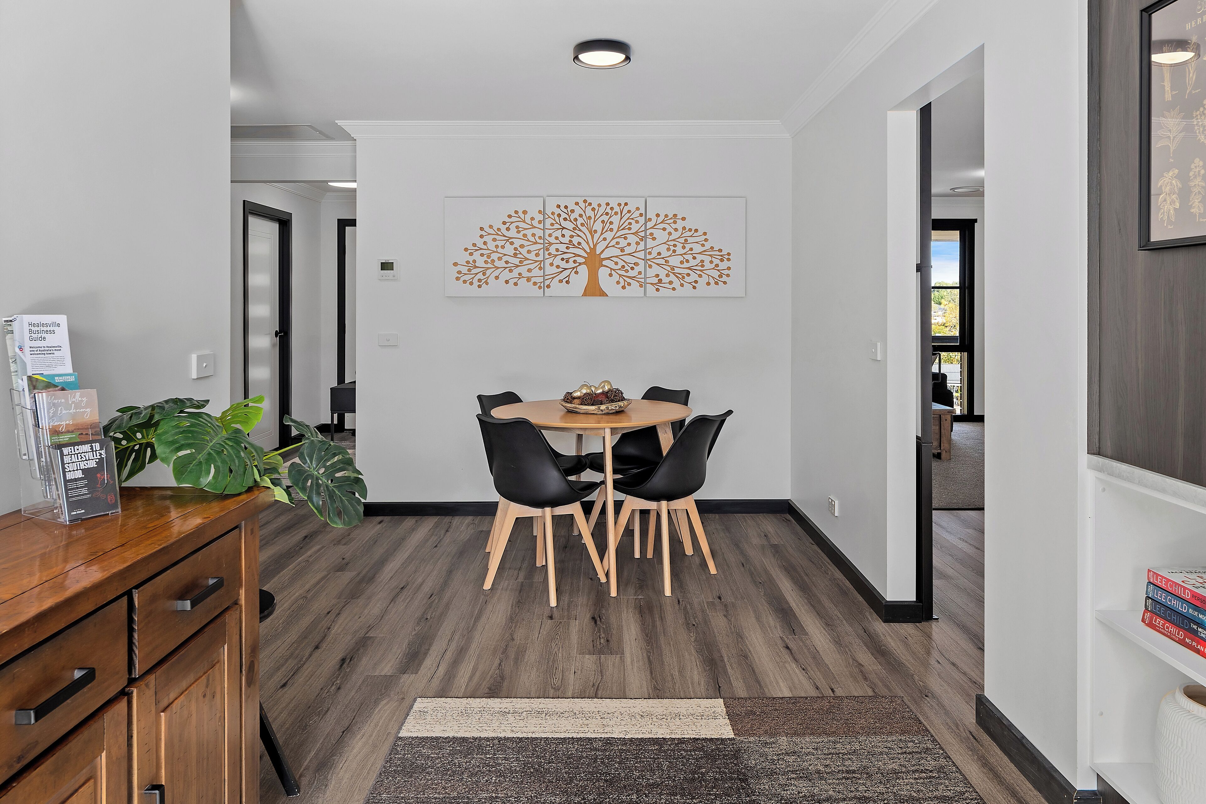 A modern dining area with a wooden table and black chairs, a large tree artwork on the wall, and a plant on the counter.