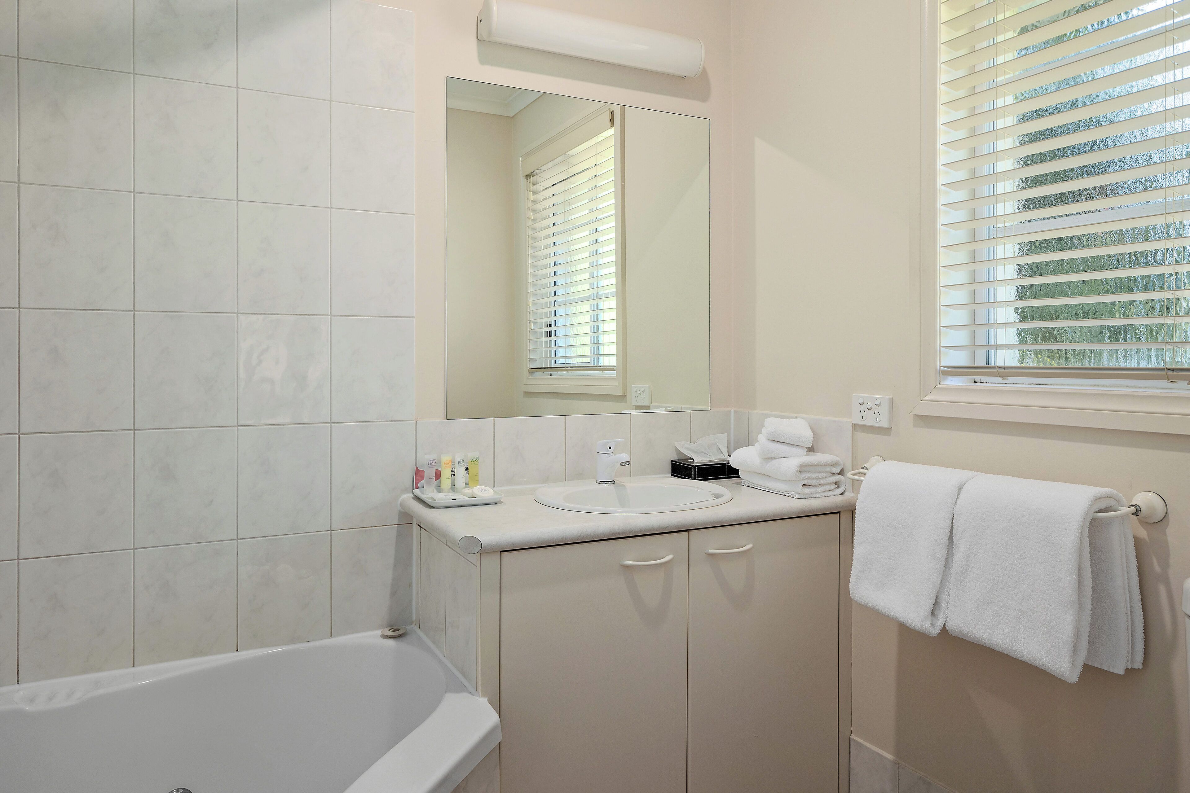 Une salle de bain avec une baignoire, un lavabo avec des articles de toilette, un porte-serviettes et une fenêtre avec des stores.