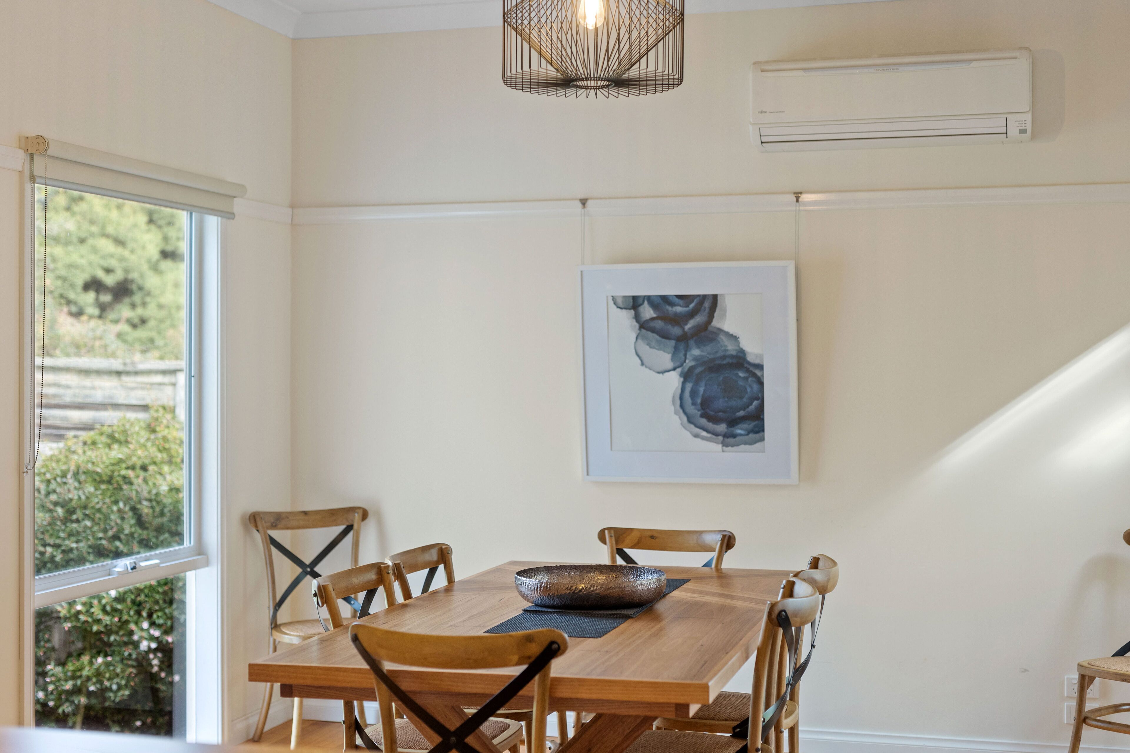 A dining area with a wooden table, six chairs, a decorative bowl, a framed artwork, and an air conditioning unit on the wall.