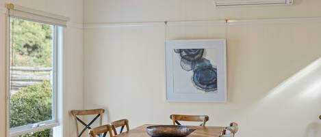 A dining area with a wooden table, six chairs, a decorative bowl, a framed artwork, and an air conditioning unit on the wall.