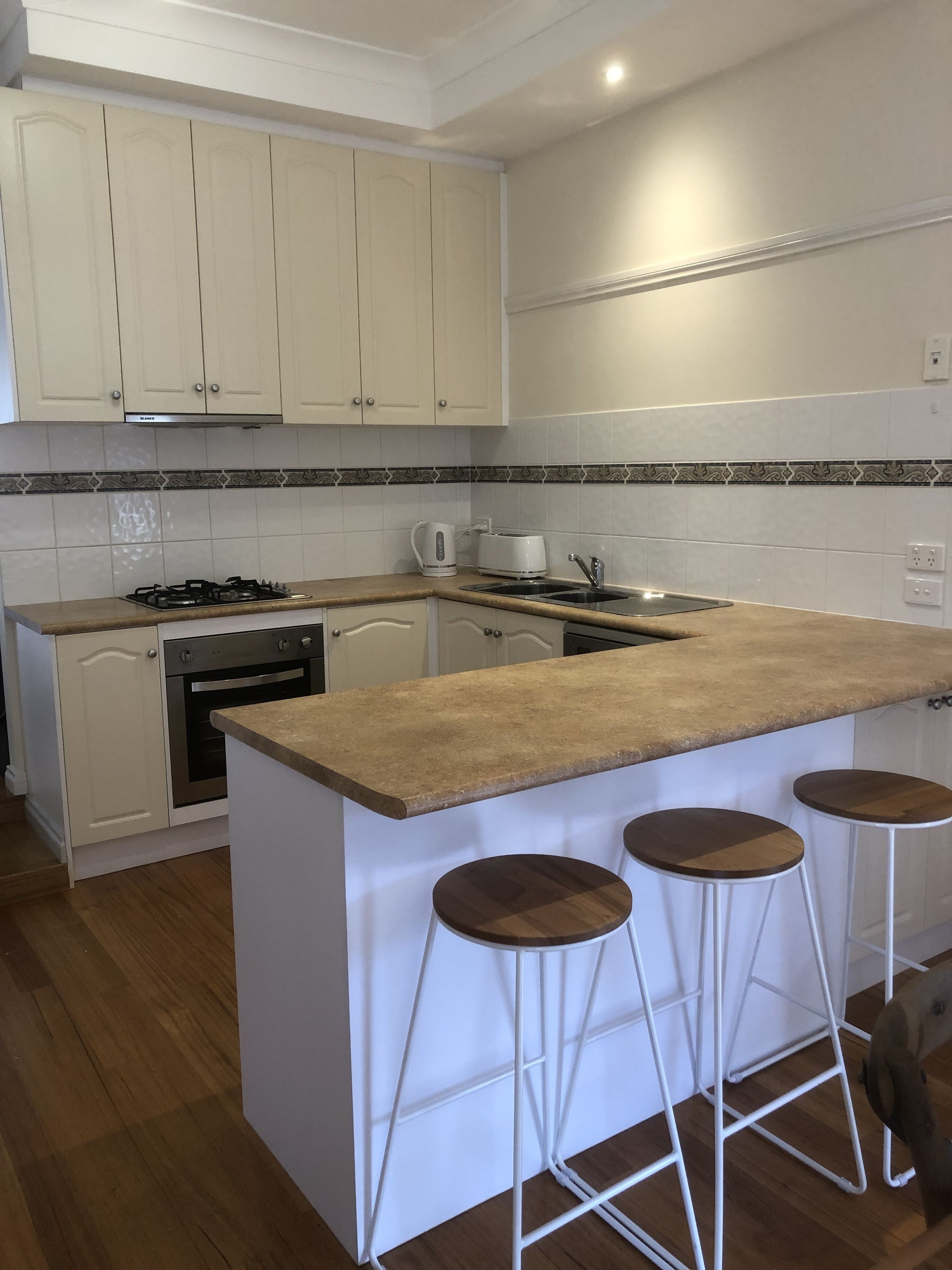 Una cocina con gabinetes blancos, estufa, horno, pileta y dos taburetes de bar.