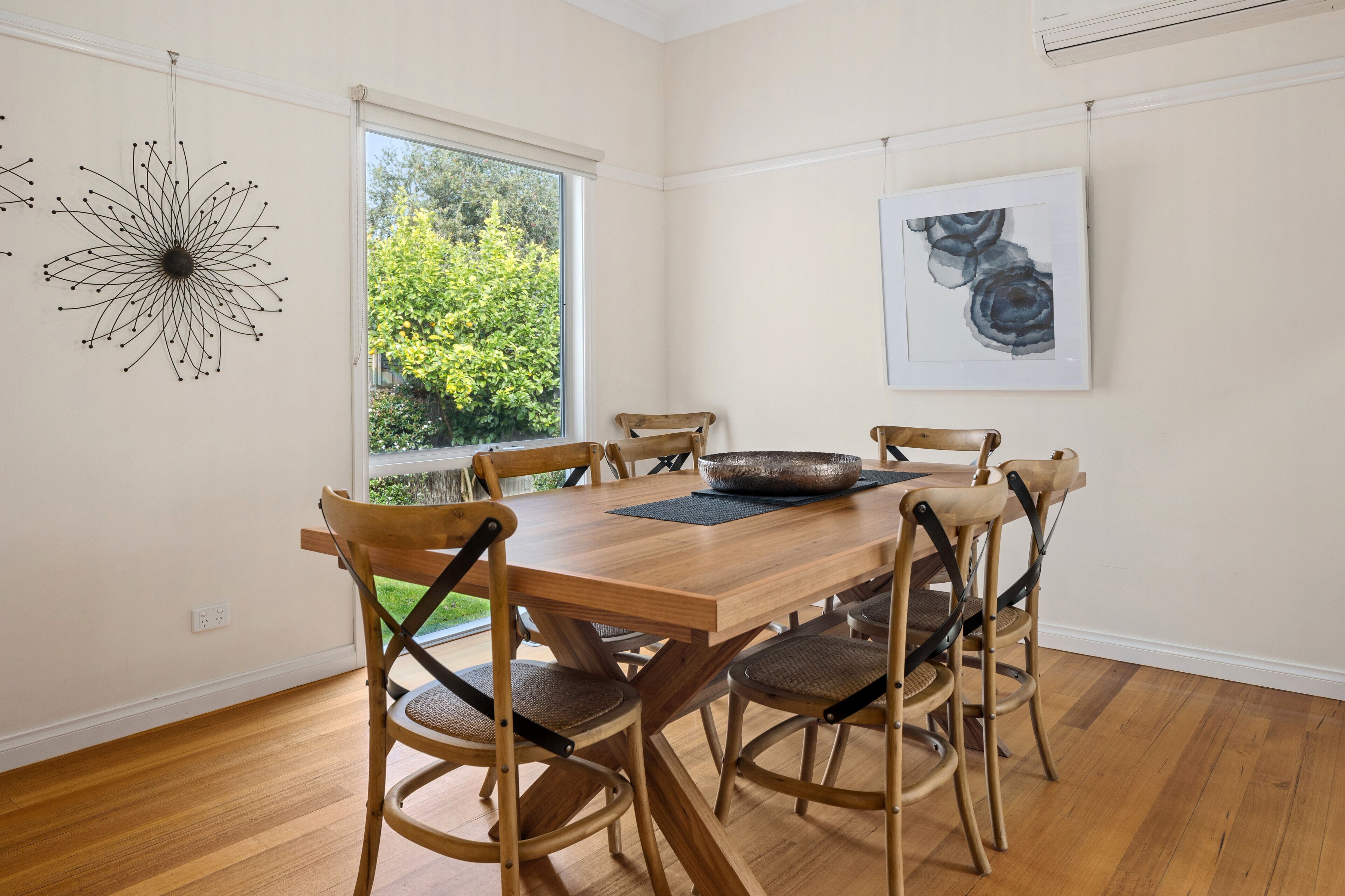 A dining area with a wooden table and chairs, a large window with a view of greenery, and a wall-mounted abstract painting.