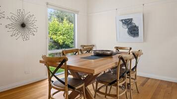A dining area with a wooden table and chairs, a large window with a view of greenery, and a wall-mounted abstract painting.