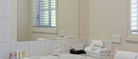 A bathroom with a white sink, mirror, and window with blinds. There are toiletries on the counter and towels stacked on the wall.