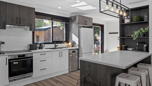 A modern kitchen with white cabinets, a built-in oven, a dishwasher, and a large island with bar stools. - Yarra Gables (Healesville)