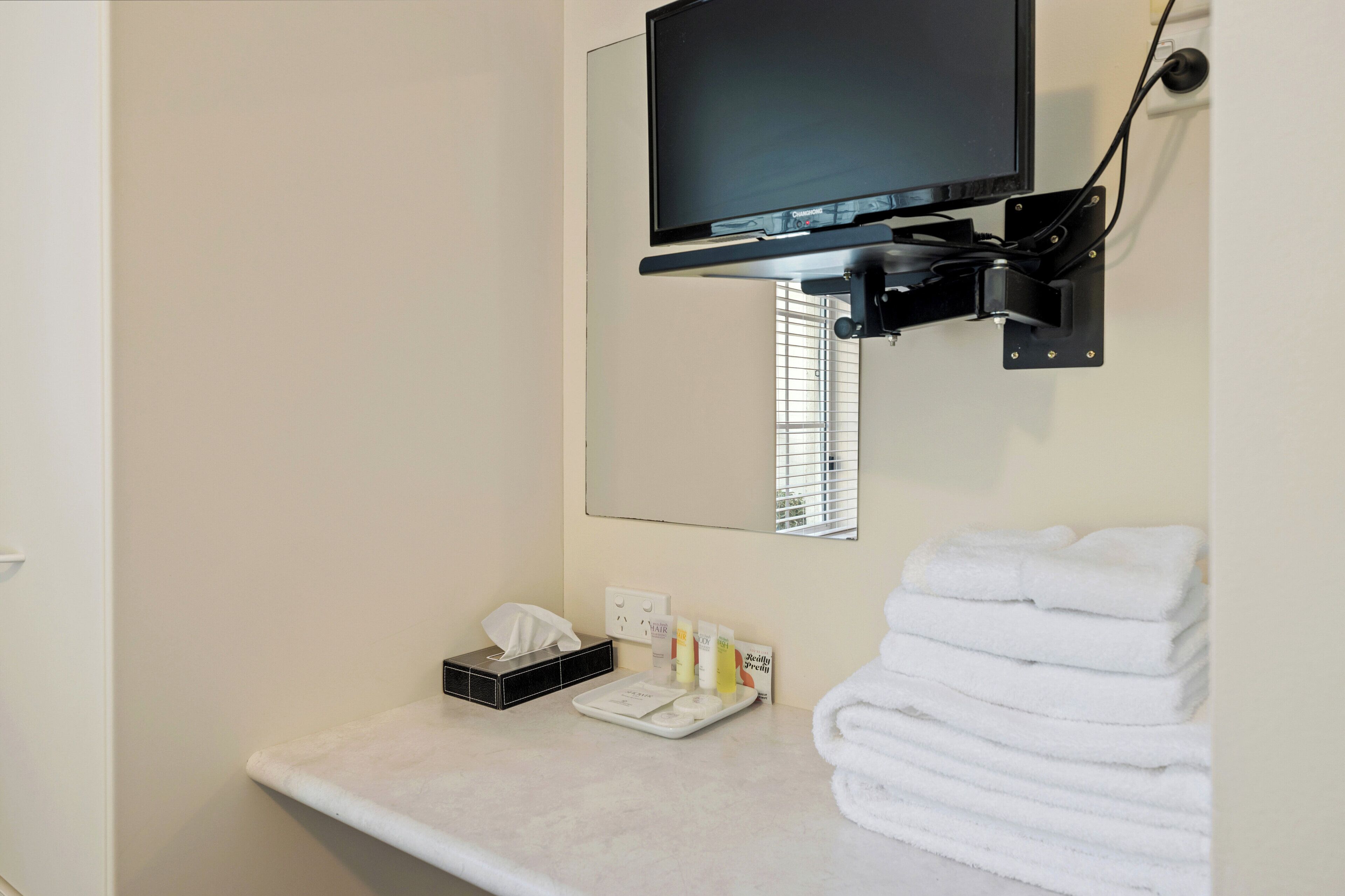 Un baño con televisión montada, toallero y un pequeño mostrador con artículos de aseo.