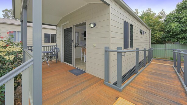 Um deck coberto com piso de madeira, uma mesa pequena e cadeiras, e uma porta que dá para um espaço interno.