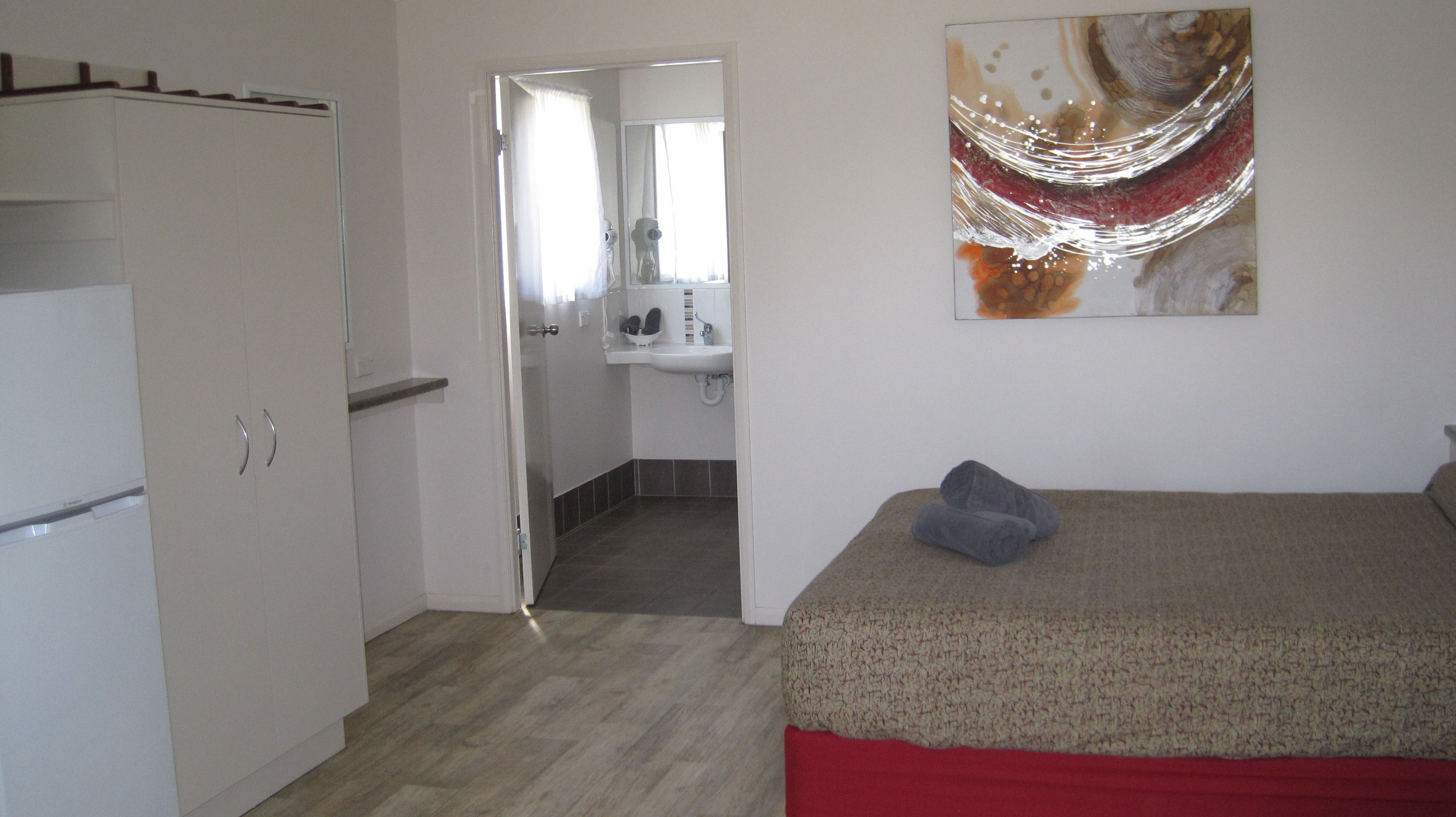 A bedroom with a bed, a red blanket, a grey pillow, a white wardrobe, a bathroom with a sink and mirror, and a painting on the wall.