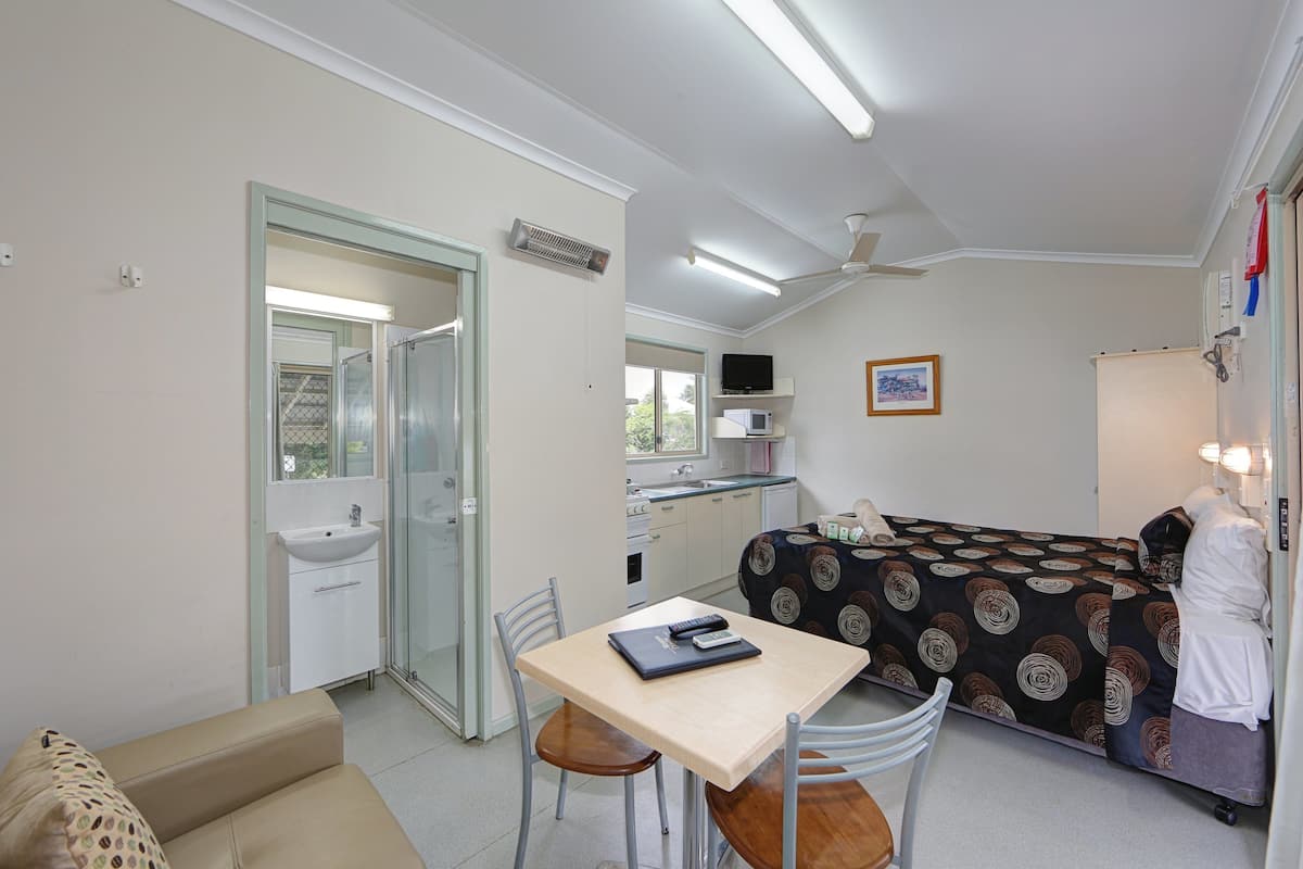 A compact hotel room with a bed, a small table with a notebook, a chair, a sink, and a television.
