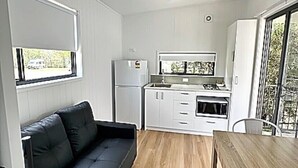 Uma cozinha compacta com armários brancos, uma geladeira e um fogão. Há um sofá preto e uma mesa de jantar de madeira com cadeiras.