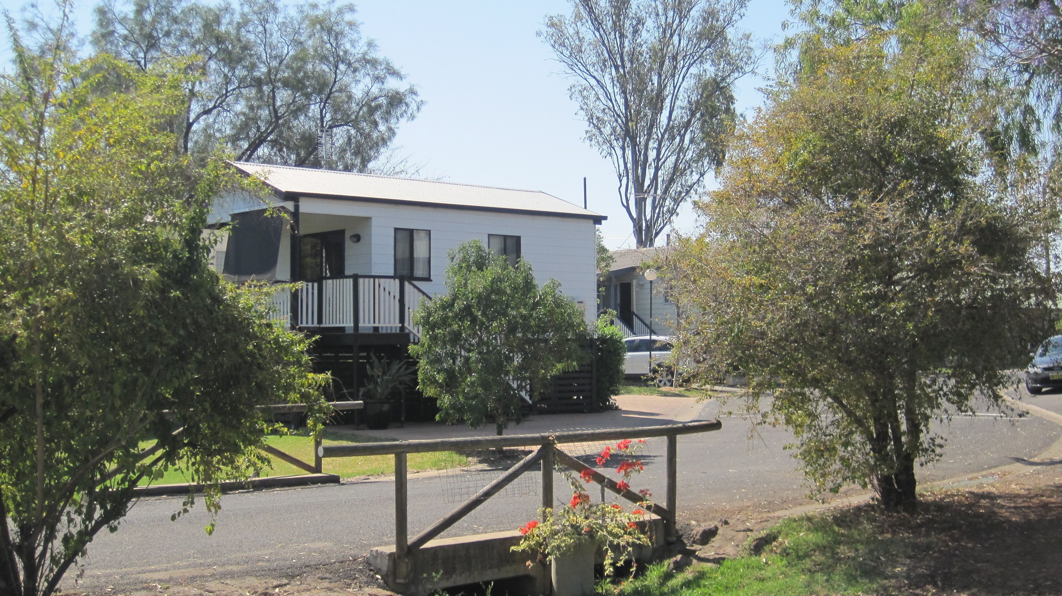 A mobile home with a balcony, surrounded by trees and a fenced area with flowers.