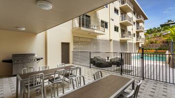 A patio area with a dining table and chairs, a grill, and a view of a building with balconies and a swimming pool.