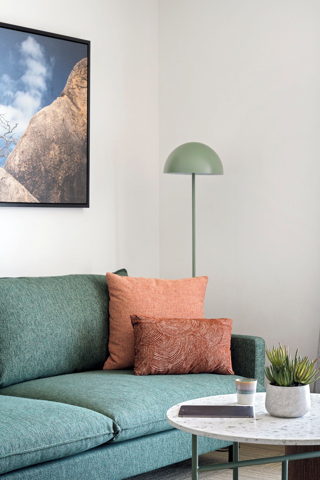 Un sofá azul turquesa con cojines estampados, una lámpara de pie verde, una mesita redonda con un libro y una planta en maceta, y un cuadro en la pared.