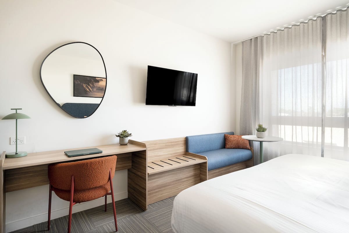 Ein modernes Hotelzimmer mit einem Holztisch, einem blauen Sofa, einem Bett, einem Fernseher und einem großen Fenster mit Vorhängen.