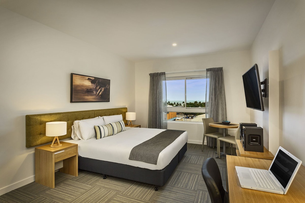 Une chambre d’hôtel comprenant un lit, des tables de chevet, un bureau avec un ordinateur portable, une télévision et une fenêtre donnant sur un paysage agréable.