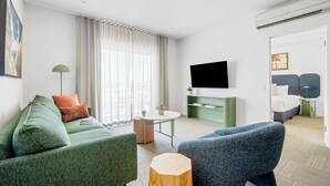 Une chambre d’hôtel moderne avec un canapé vert, un fauteuil bleu, une table ronde et un téléviseur à écran plat fixé au mur.