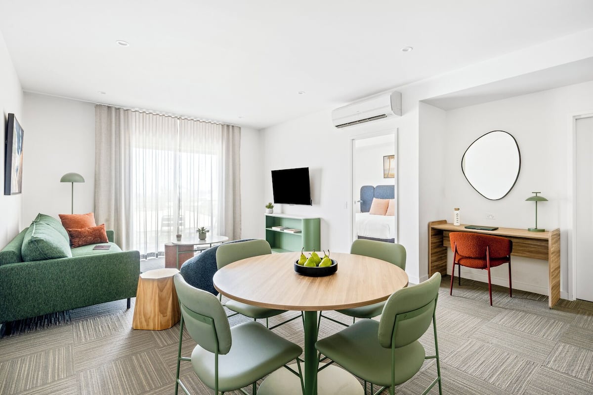 Ein modernes, helles Wohnzimmer mit einem grünen Sofa, einem runden Holzesstisch und einem an der Wand befestigten Fernseher.