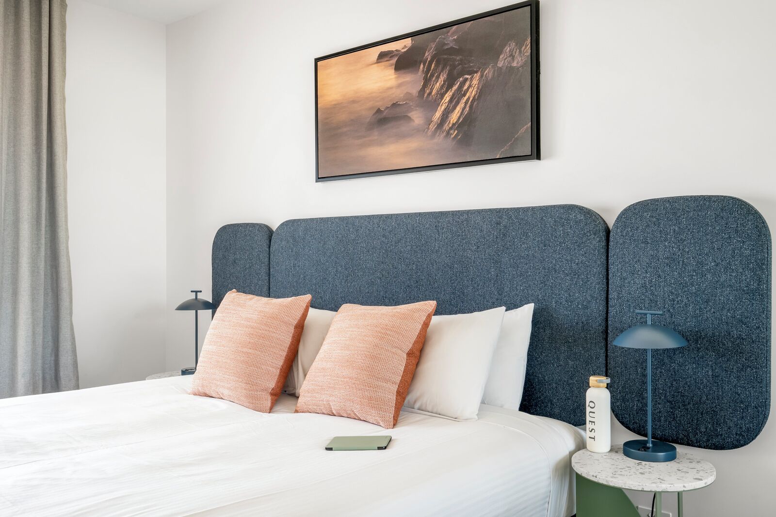 Une chambre d’hôtel moderne avec un grand lit, deux oreillers, une table de chevet avec une lampe, un tableau encadré au mur et une petite table avec une bouteille.