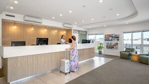Un espace d’accueil d’hôtel au design moderne, comprenant un comptoir de réception portant l’inscription « QUEST », des unités de climatisation et un coin salon offrant une vue sur l’extérieur.