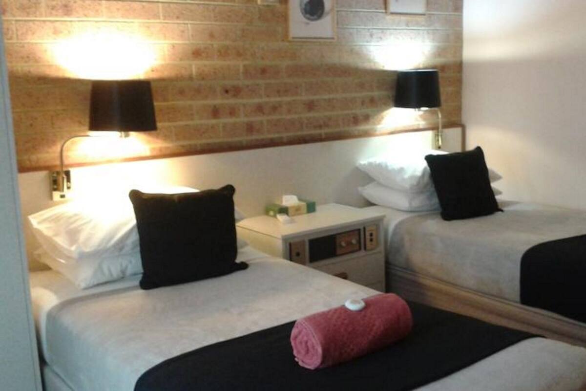 A hotel room with two beds, each with a black pillow and a rolled towel. There are wall-mounted lamps and a small table with a lamp between the beds.