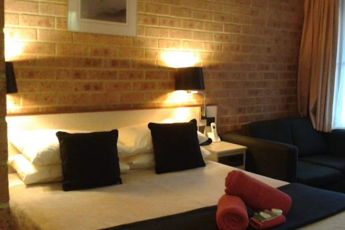 A hotel room with a brick wall, a bed with black pillows, a nightstand, a red towel, and a dark-colored sofa.
