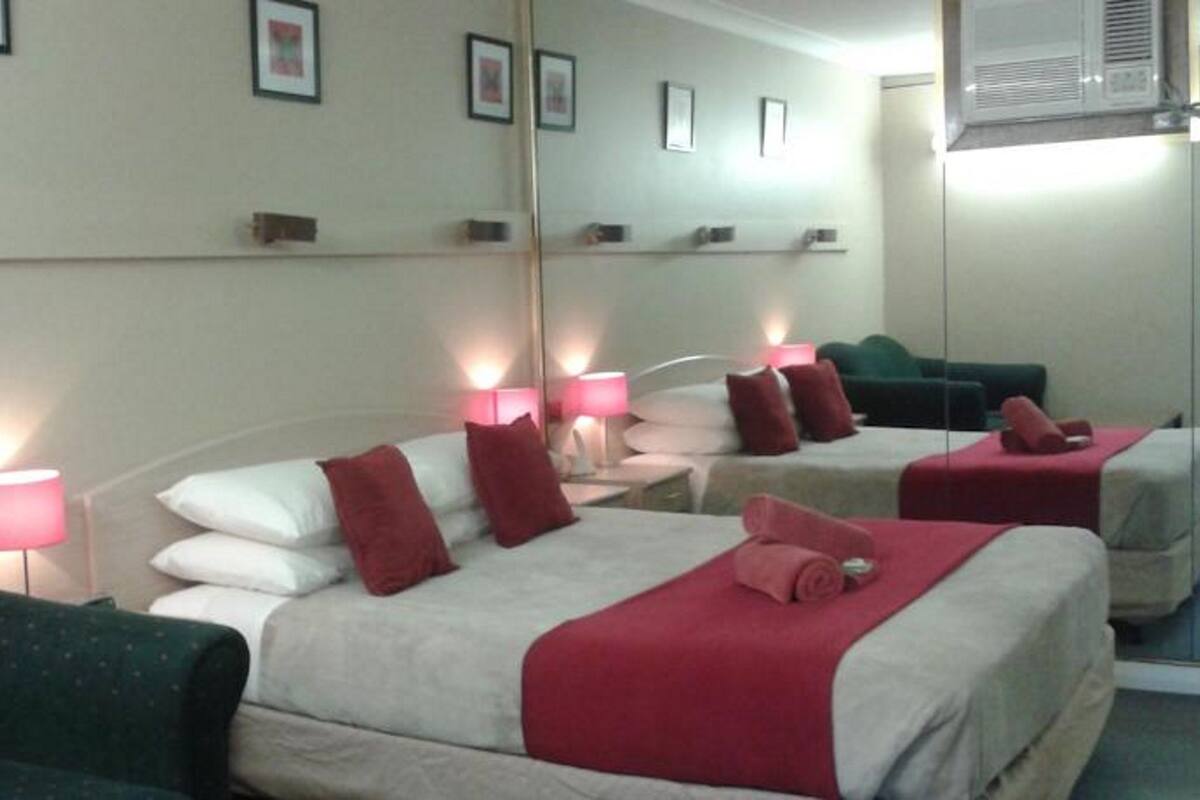 A hotel room with two beds, each with red and white bedding, a green armchair, bedside lamps, and a mirror on the wall.