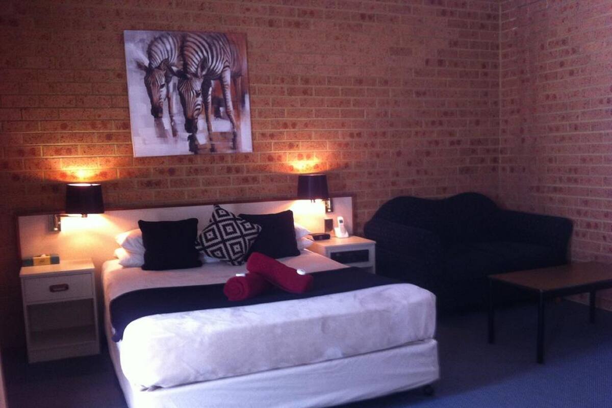 A hotel room with a brick wall, a bed with white bedding, two bedside tables with lamps, a black sofa, and a zebra painting on the wall.