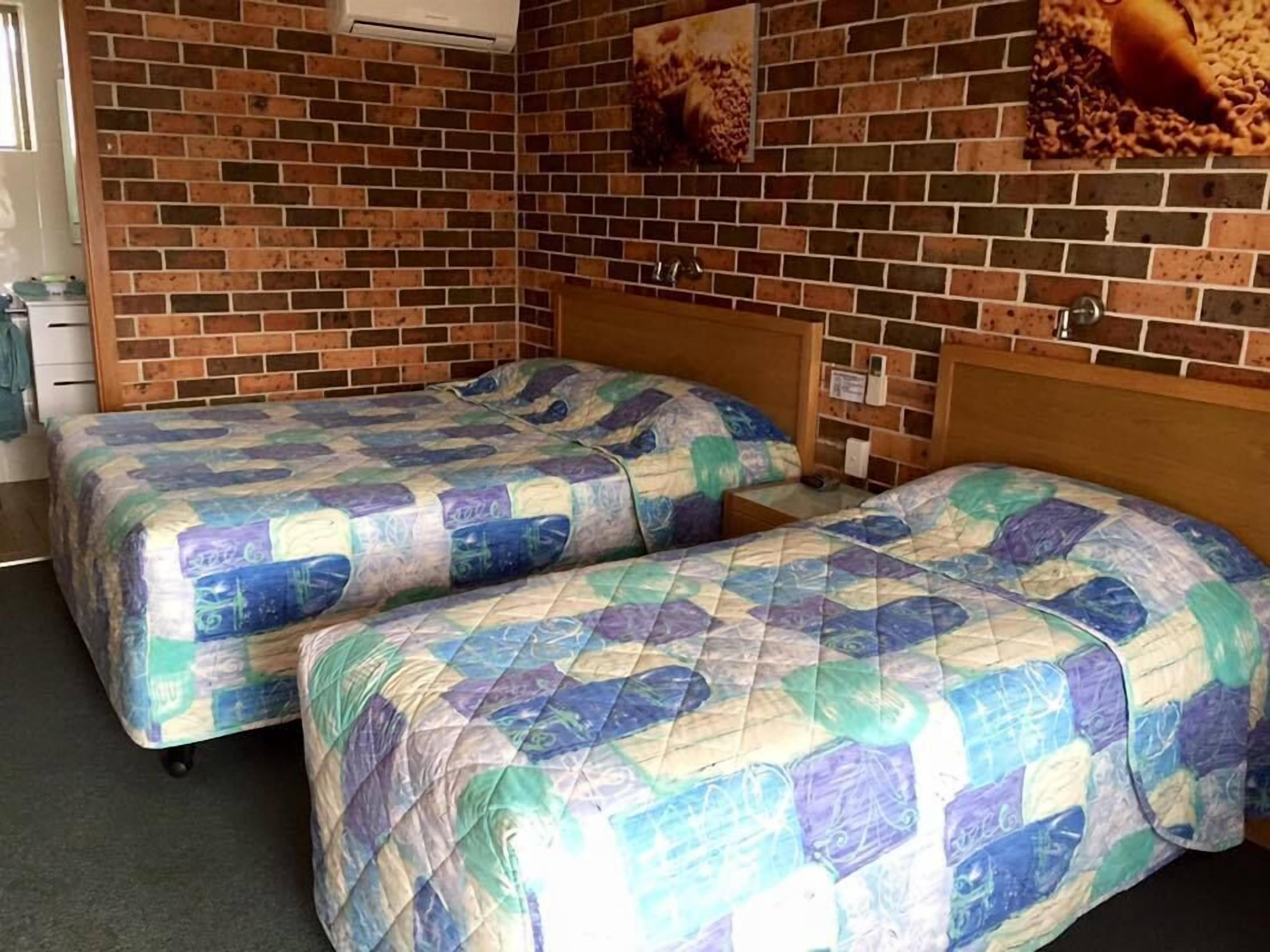 A room with two beds, each with a quilted bedspread, a brick wall, and a picture of food on the wall.