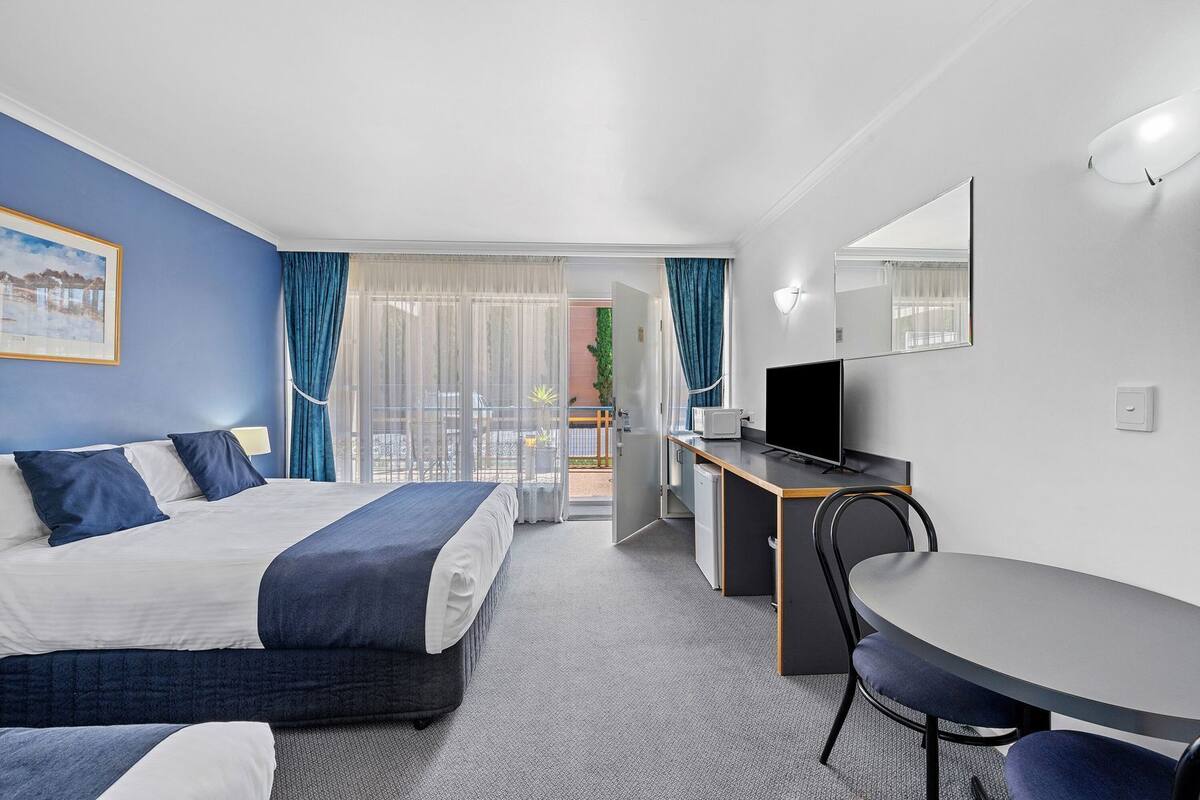 A hotel room with a bed, a desk with a TV, a chair, and a balcony with curtains.