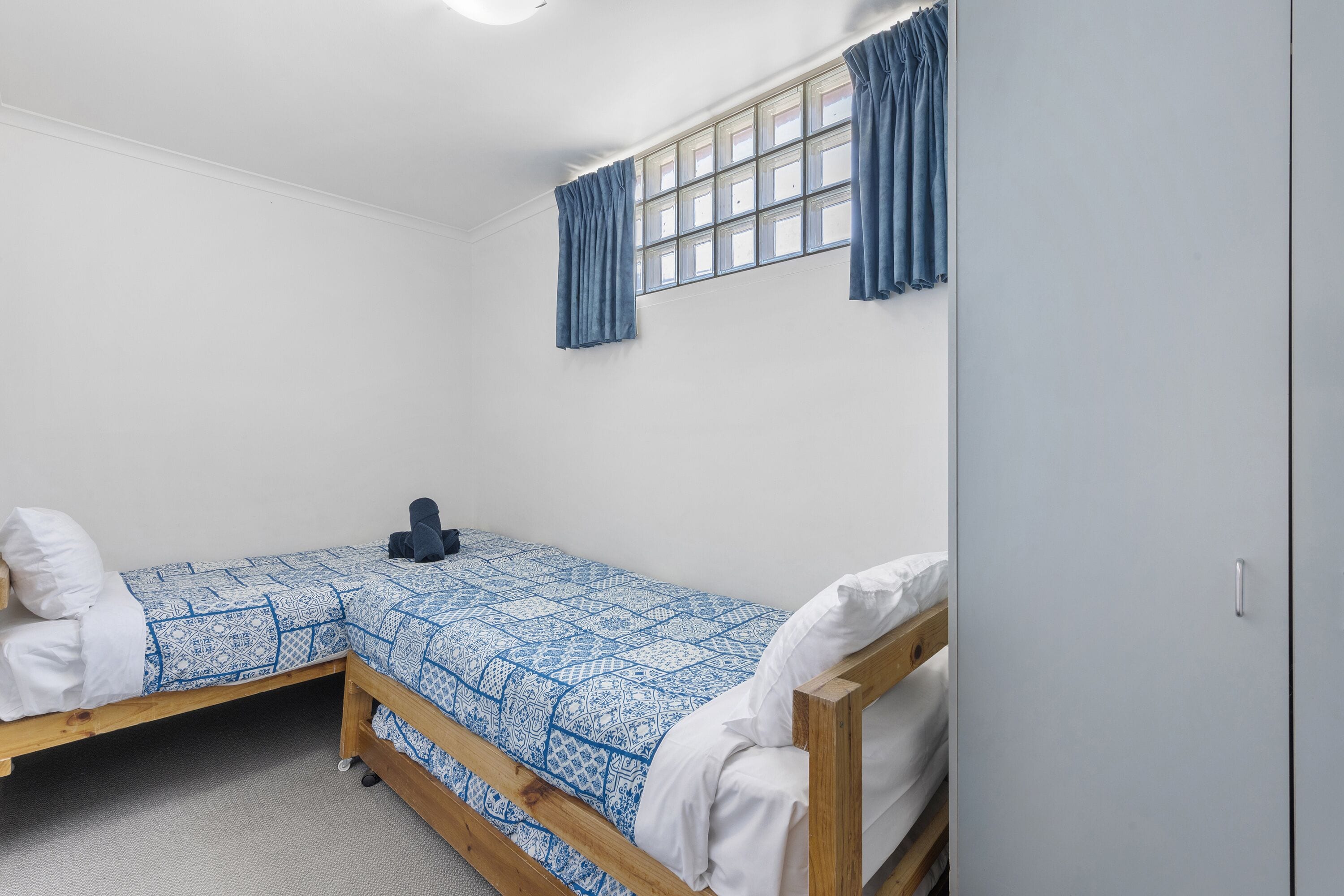 A small, single-bed room with a wooden frame, a patterned blue and white bedspread, and a window with blue curtains.