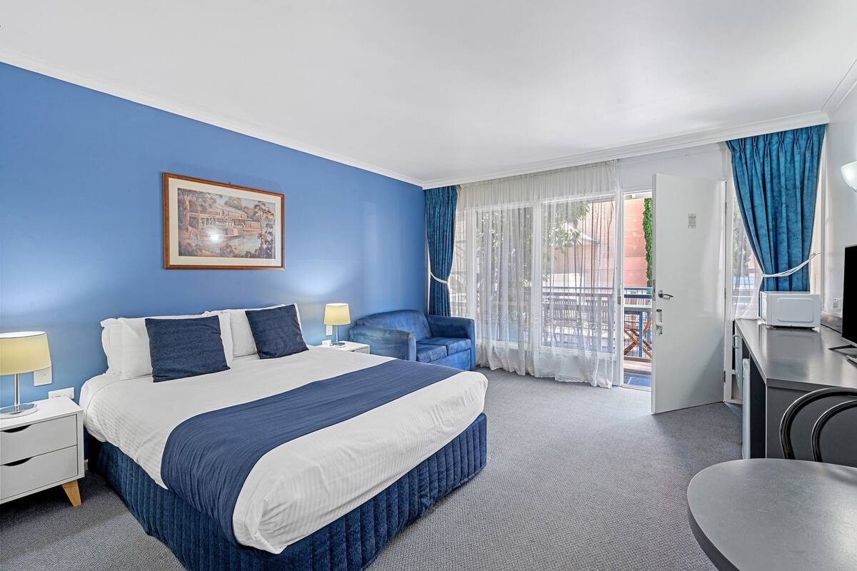 A hotel room with a bed, a desk, a chair, a nightstand, a lamp, a picture on the wall, and a view of the outside.