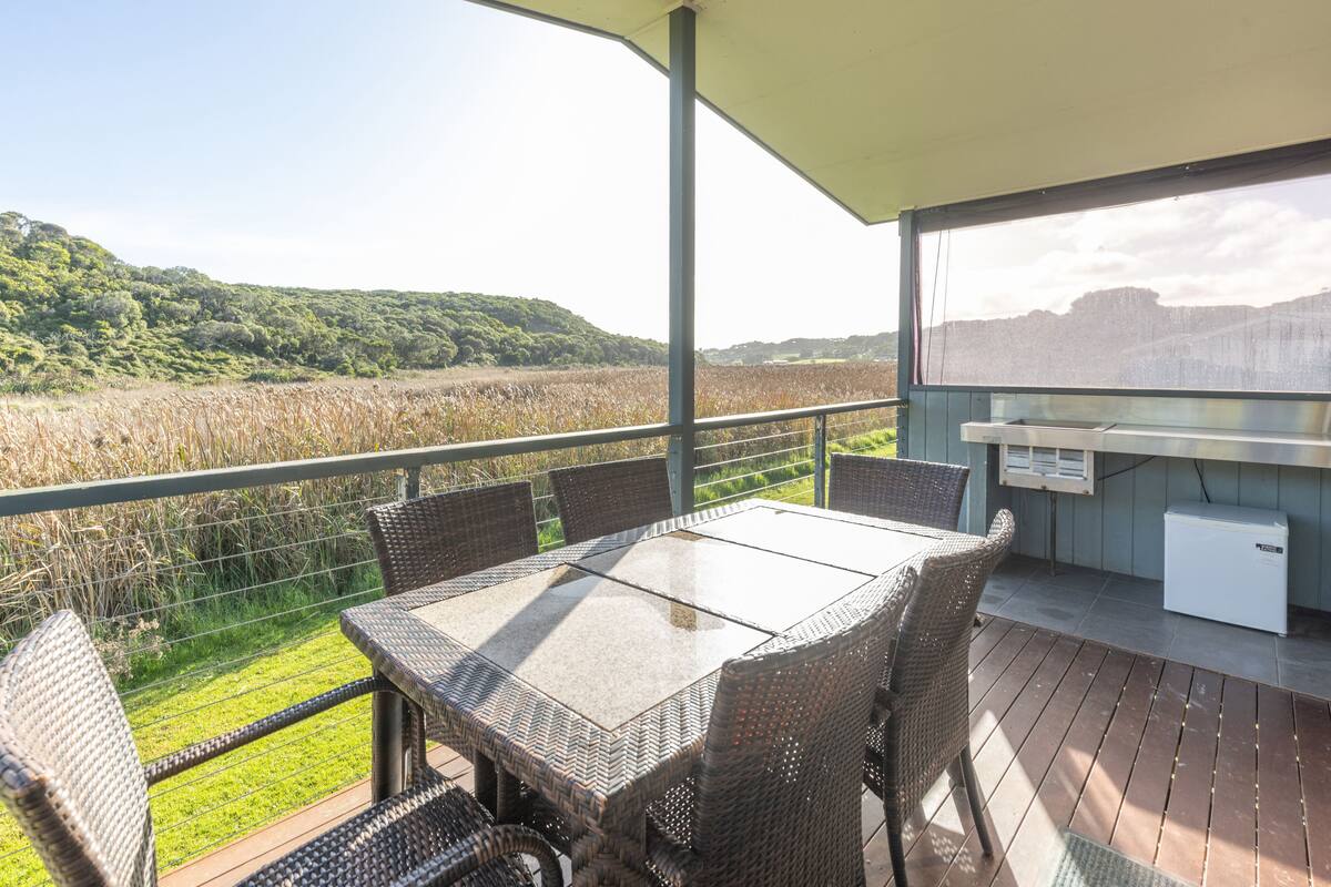 Un espacio al aire libre cubierto con mesa y sillas, un refrigerador y vistas a un campo y colinas.
