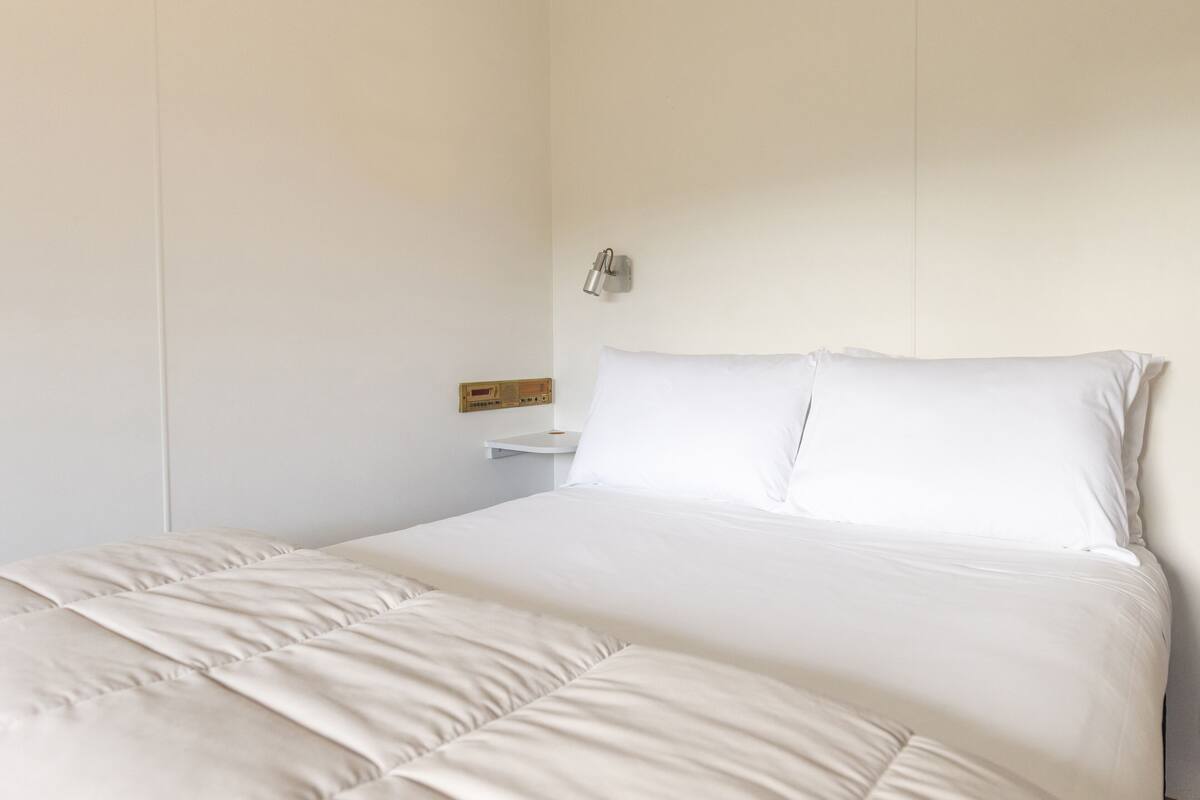Una cama bien hecha con edredón blanco y almohadas, una mesita de noche con una radio y una lámpara fijada a la pared.