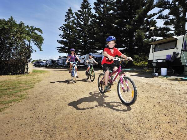 Três crianças andando de bicicleta em uma trilha de terra, com uma floresta e veículos tipo RV ao fundo.