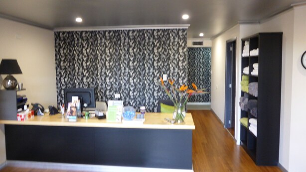 A modern reception desk with a dark wood finish, a patterned wallpaper backdrop, and a vase with flowers. - Tanoshii Spa & The Edge Spa Stay (Goonellabah)