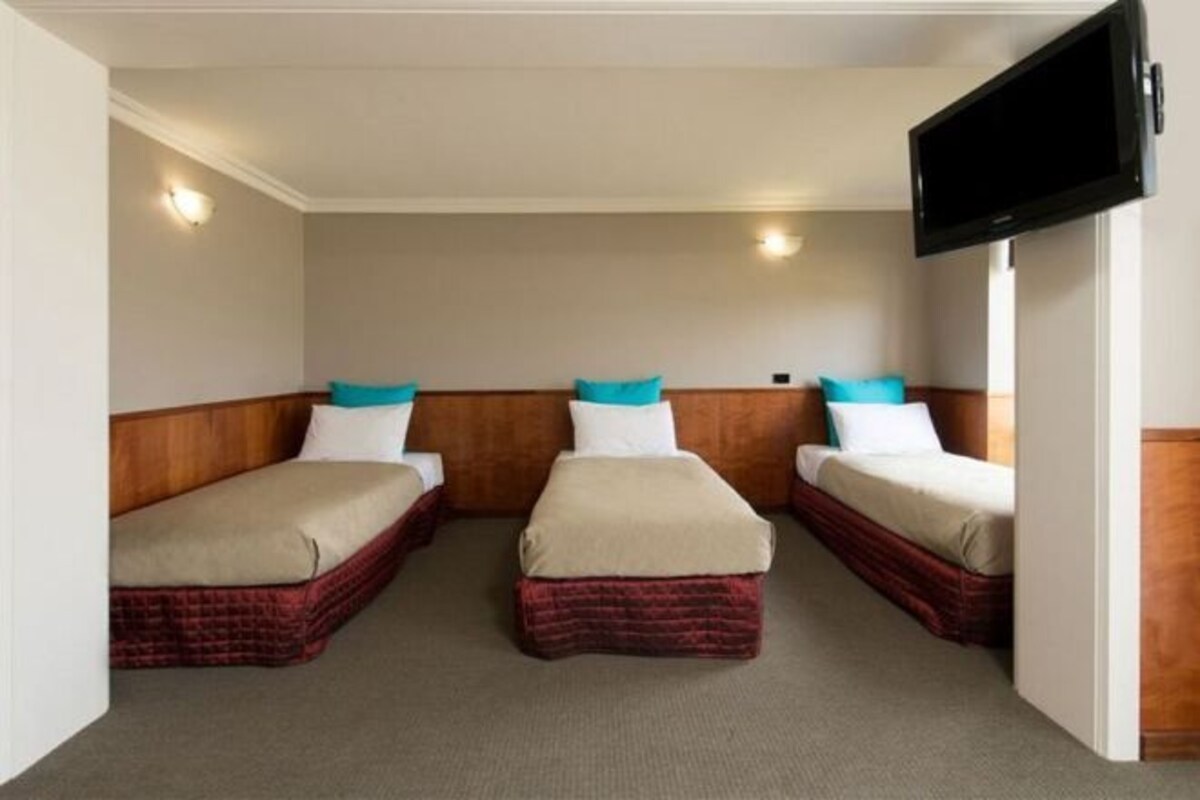 A hotel room with two single beds, each with a wooden headboard and a red bed skirt. There is a flat-screen TV mounted on the wall to the right.