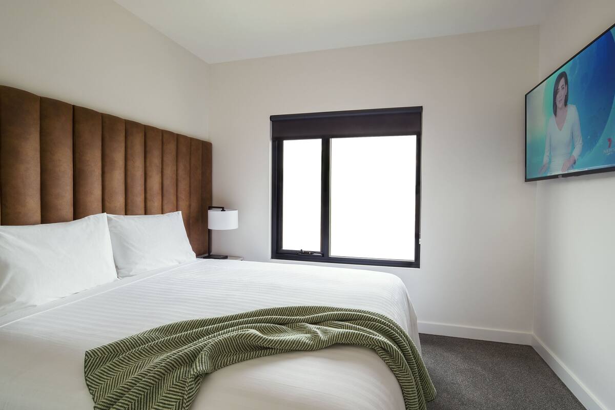 A hotel room with a large bed, a window, a wall-mounted TV, and a painting.