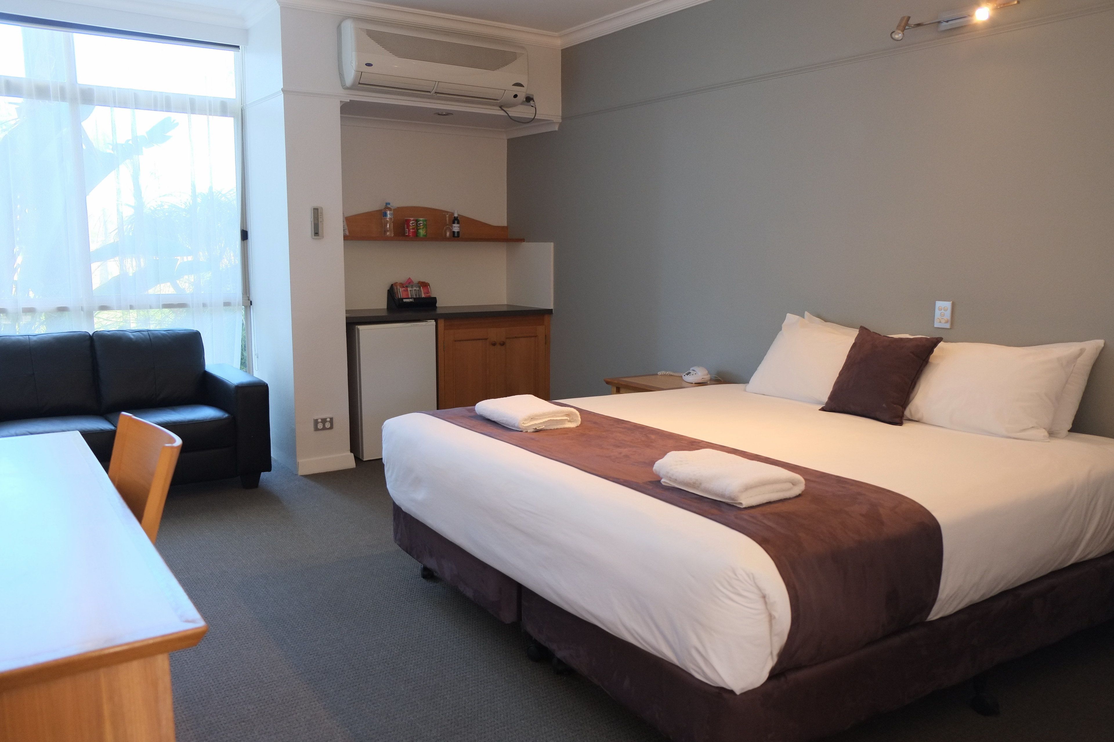 A hotel room with a bed, a desk, a chair, a kitchenette, and a window.