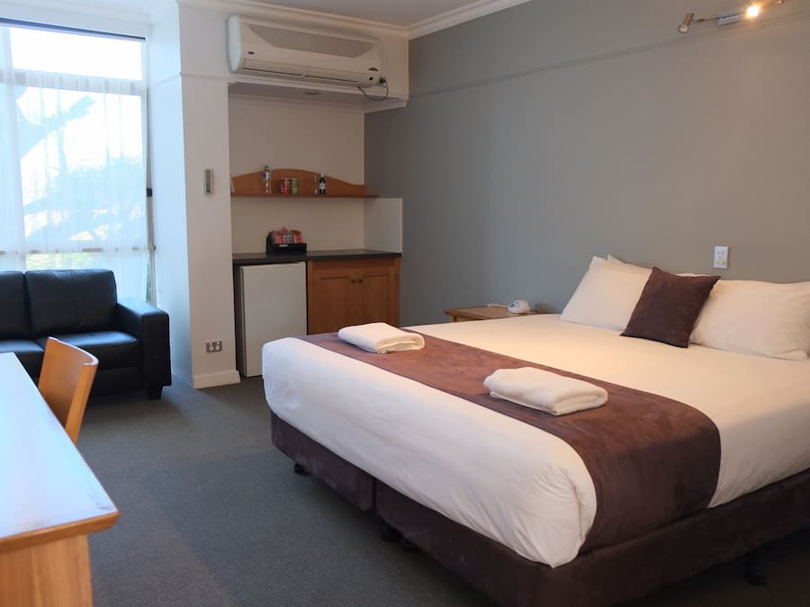 A hotel room with a bed, a desk, a chair, a kitchenette, and a window.