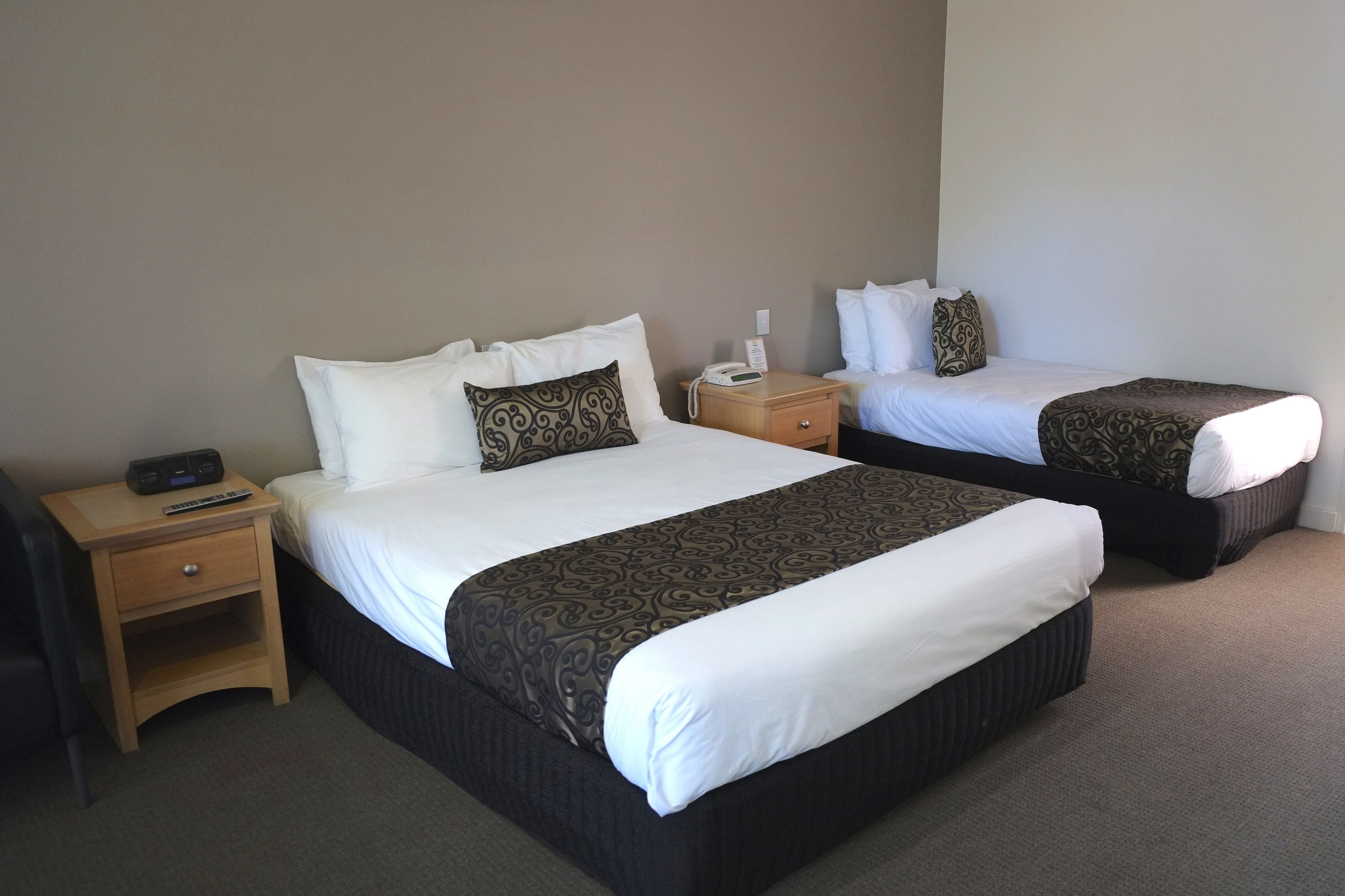 A hotel room with two beds, each with a patterned bedspread and white pillows. There is a wooden nightstand with a phone and a remote control.