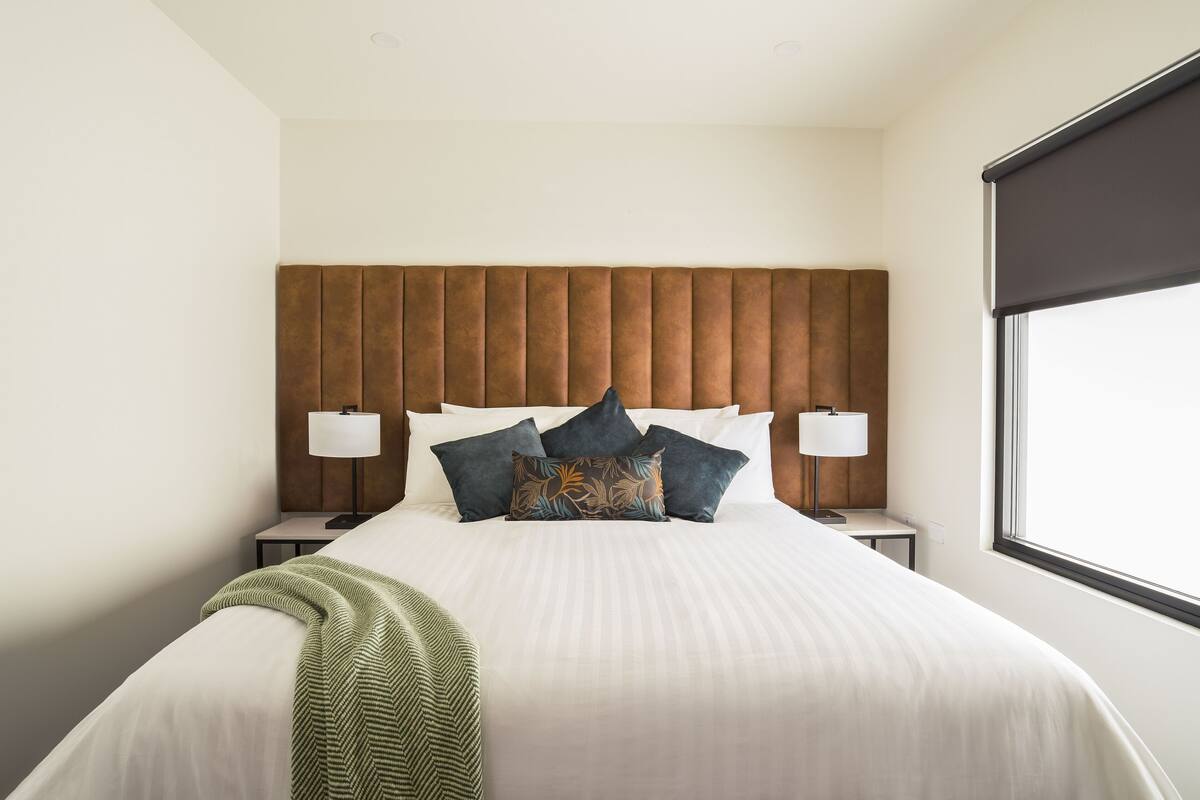 A neatly made bed with white linens and decorative pillows, a wooden headboard, bedside lamps, and a window with a view.