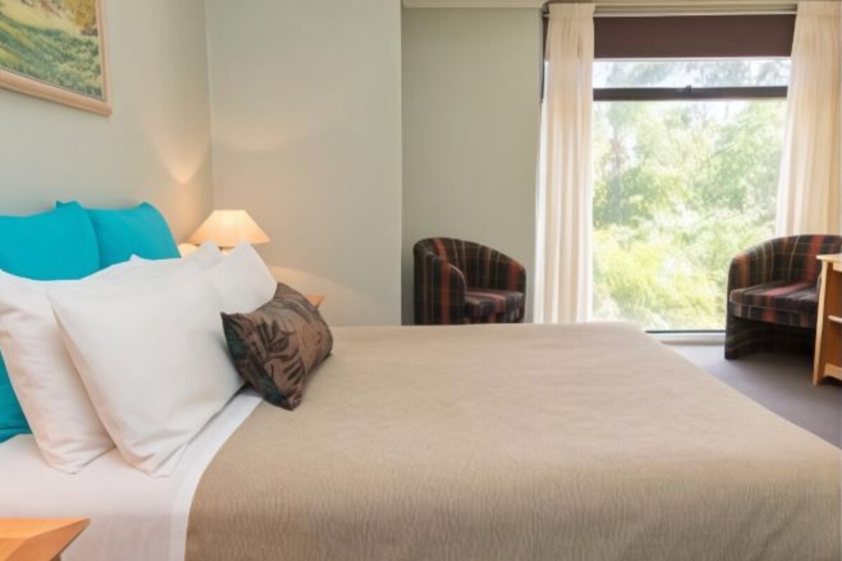 A hotel room with a bed, a nightstand, a lamp, two chairs, and a window with curtains.