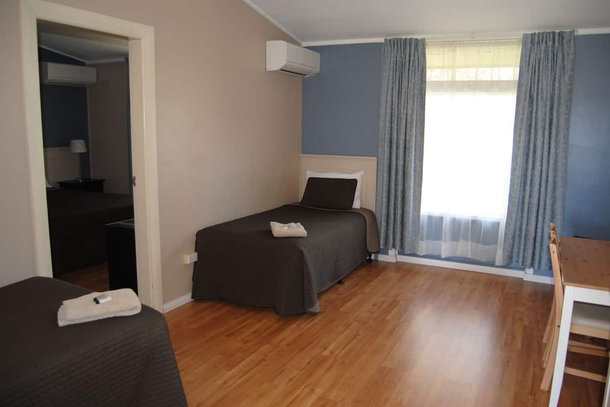 A bedroom with a bed, a desk, and a chair. There is a window with curtains, an air conditioning unit, and a door leading to another room.