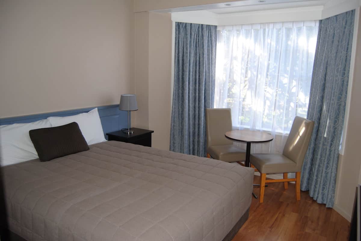 A hotel room with a large bed, a small table, two chairs, a lamp, and a window with curtains.
