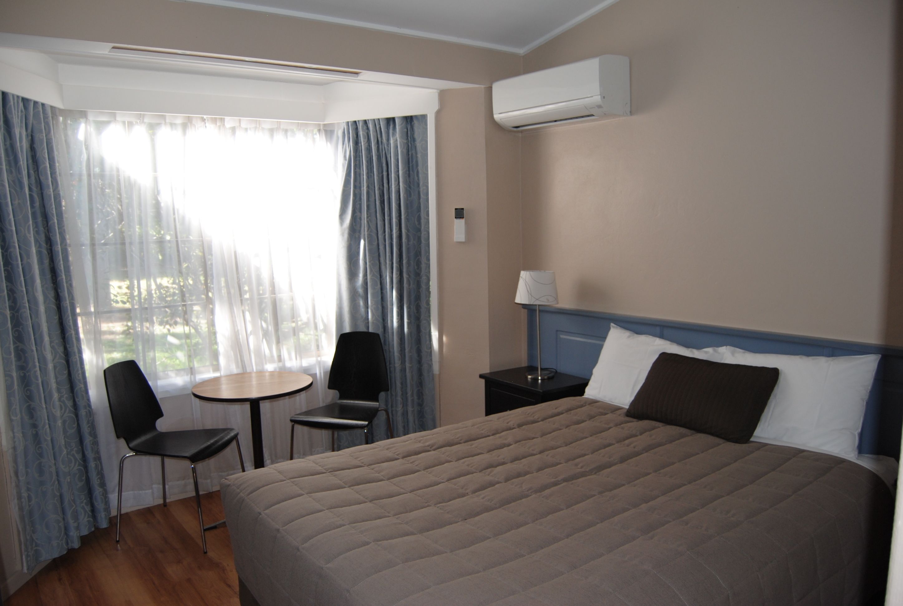A hotel room with a bed, a small table, two chairs, a lamp, and an air conditioning unit.