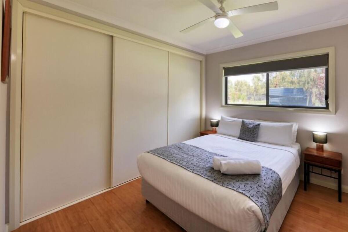 A bedroom with a bed, bedside tables, a ceiling fan, and a window with a view of trees.