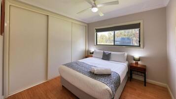 Ein Schlafzimmer mit Bett, Nachttischen, Deckenventilator und Fenster mit Blick auf Bäume.
