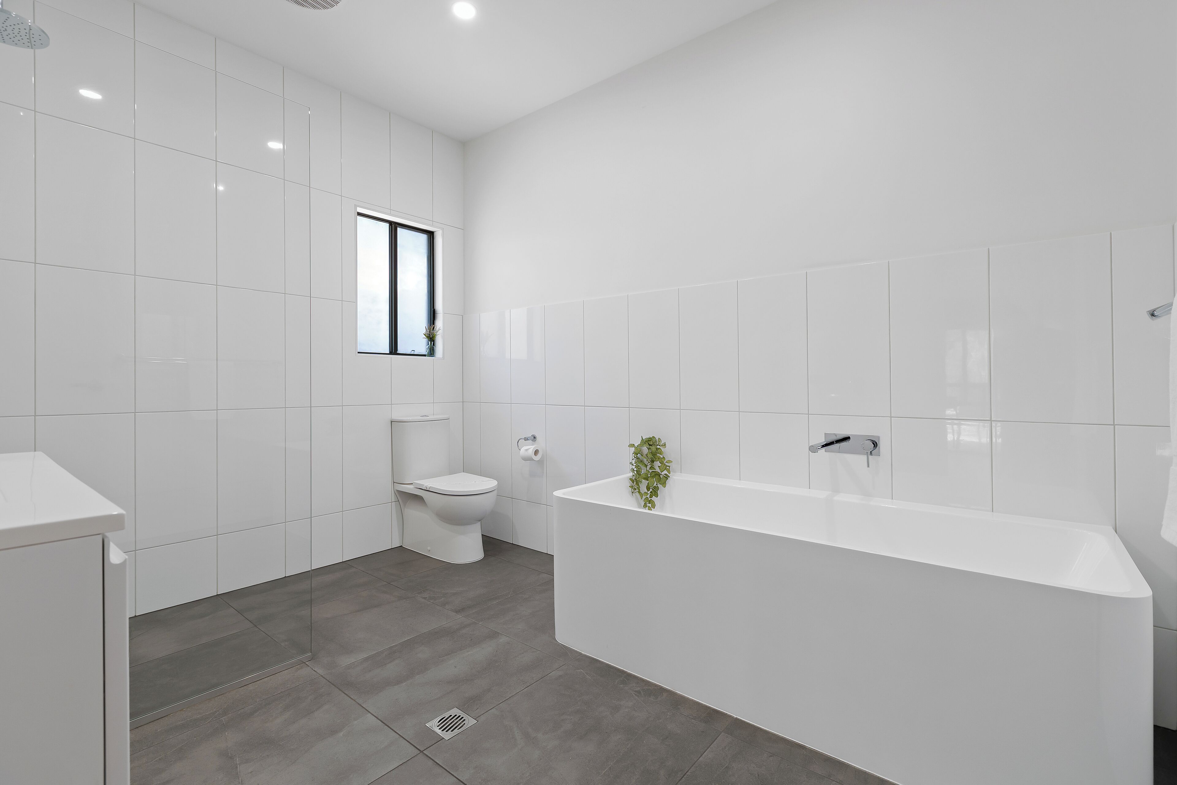 A modern bathroom with white tiles, a toilet, a bathtub, and a window.